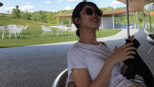 A photo of a girl wearing a white t-shirt and sunglasses, sitting in an open terrace. In the background there is a forest, some green grass, some white tables and chairs.