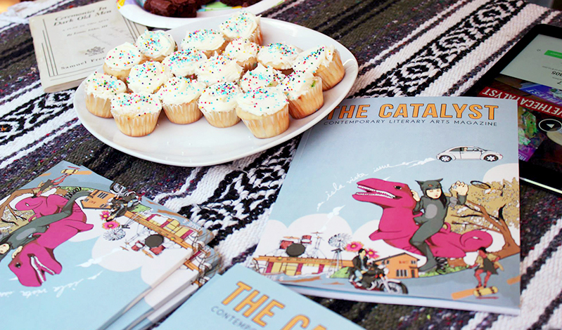 A photo of a plate filled with white cup cakes and some comic books. Each comic book has a drawing of a pink dinosaur ridden by a girl in a cat costume, a white car flying a man on a motorcycle and a house in the background. The title of the comic book: The Catalyst.