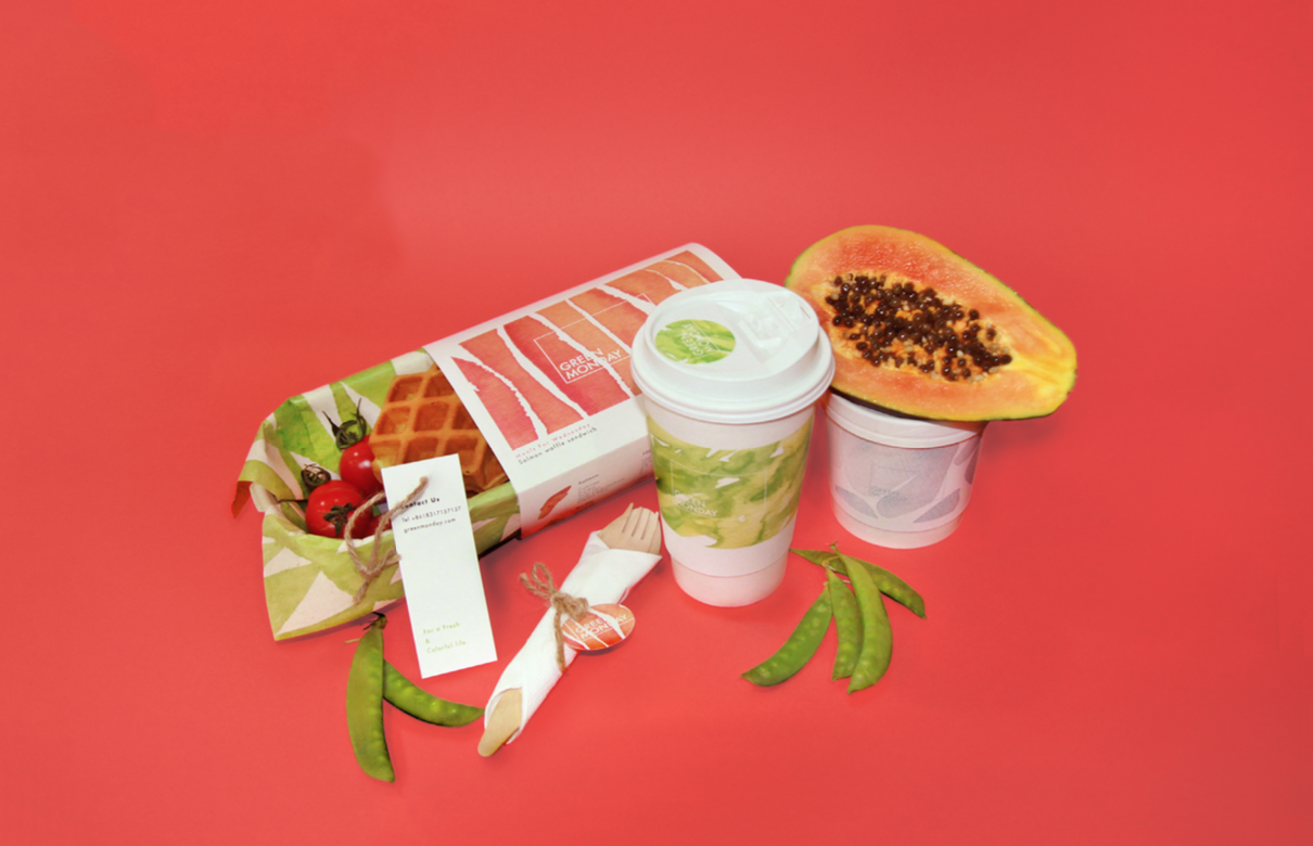 Different green vegetables, a plastic cup, wrapped cutlery, plastic jar, avocado and some packed waffles on a red background.