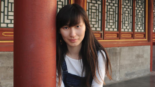 A girl sitting near a red, round stone pillar in front of a building.