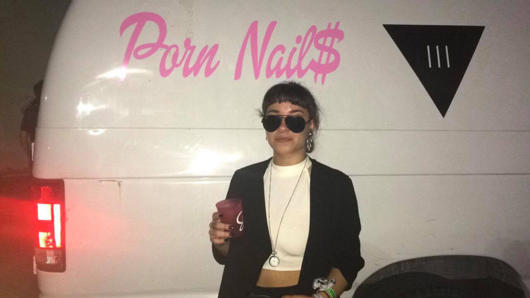A photo of a girl waring sunglasses while standing near a white wan it some pink text on it.