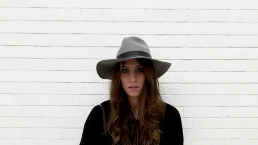A photo of a woman wearing a hat and a black dress, standing in front of a white brick wall.