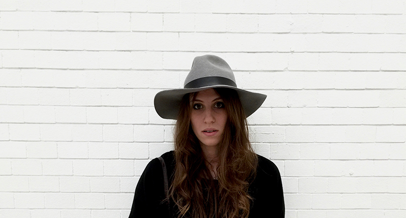 A photo of a woman wearing a hat and a black dress, standing in front of a white brick wall.
