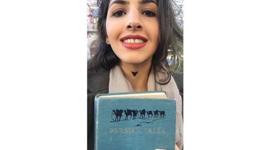A photo of a smiling girl with a blue book in her hands on which there are some camels an the text: Persian Tales.