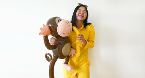 A photo of a girl wearing a yellow suit with a banana in one hand and a balloon brown monkey in another hand.