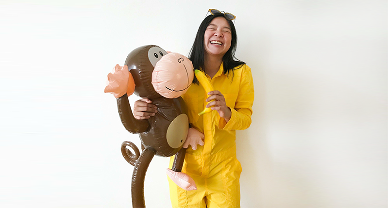 A photo of a girl wearing a yellow suit with a banana in one hand and a balloon brown monkey in another hand.