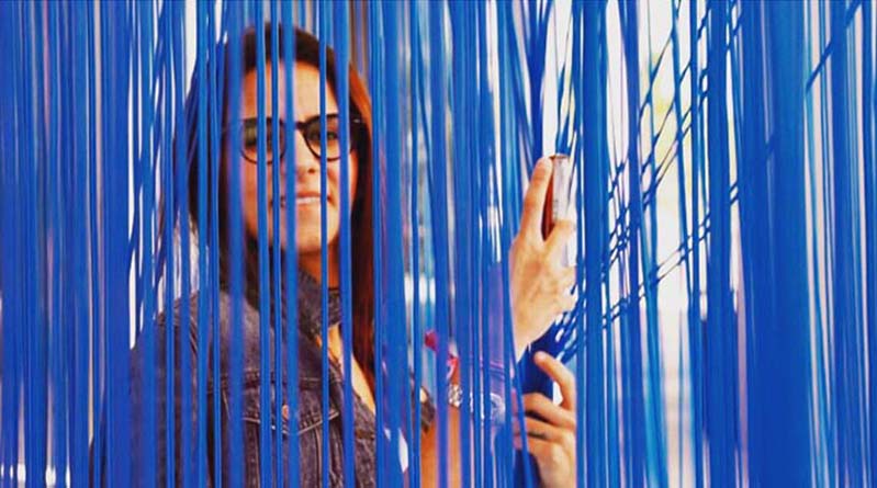 A photo of a woman wearing glasses and sitting behind a curtain with blue fringes.