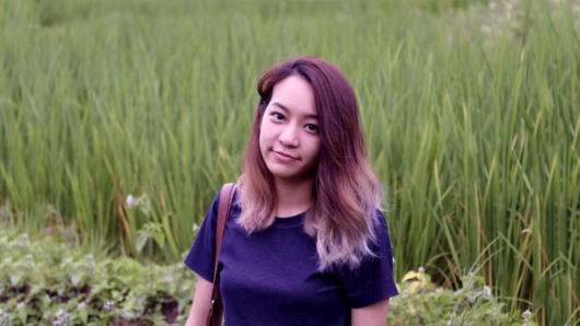 A photo of a woman wearing a blue t-shirt, standing in front of a green field.