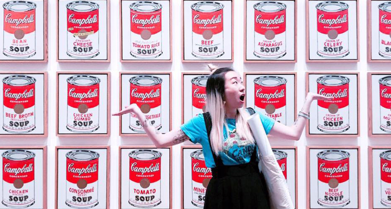 A photo of a girl wearing a blue t-shirt, black pants and caring a white bag with a paper scroll in it. The girl is in front of wall with a texture made from images of red and white tin cans.