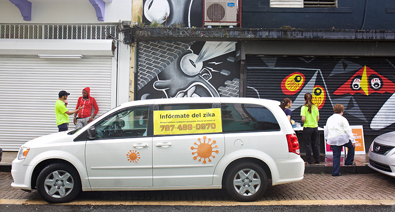 A photo of a white wan with a yellow placard with text and a phone number on it. The van has also some orange splashes on it.