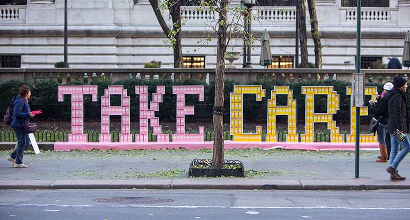 A street art setup made from plastic that depicts big letters forming the text: Take Care. Also, placed in the letters are small pink and yellow boxes.