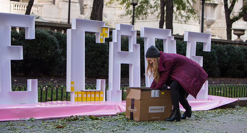 A photo of a woman taking something from a card box while standing near a pink art setup that depicts big letters in which you can place small yellow boxes. The text reads Take Care.