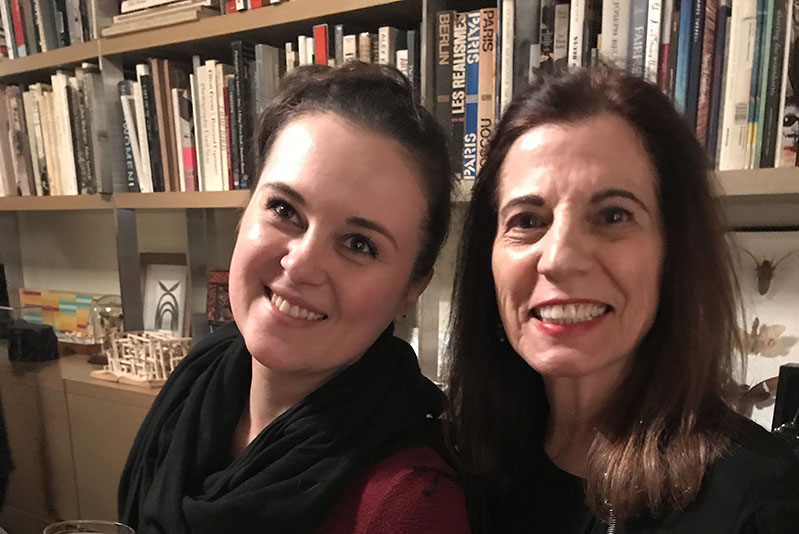 A photo of two smiling women  sitting in front of a book shelf.