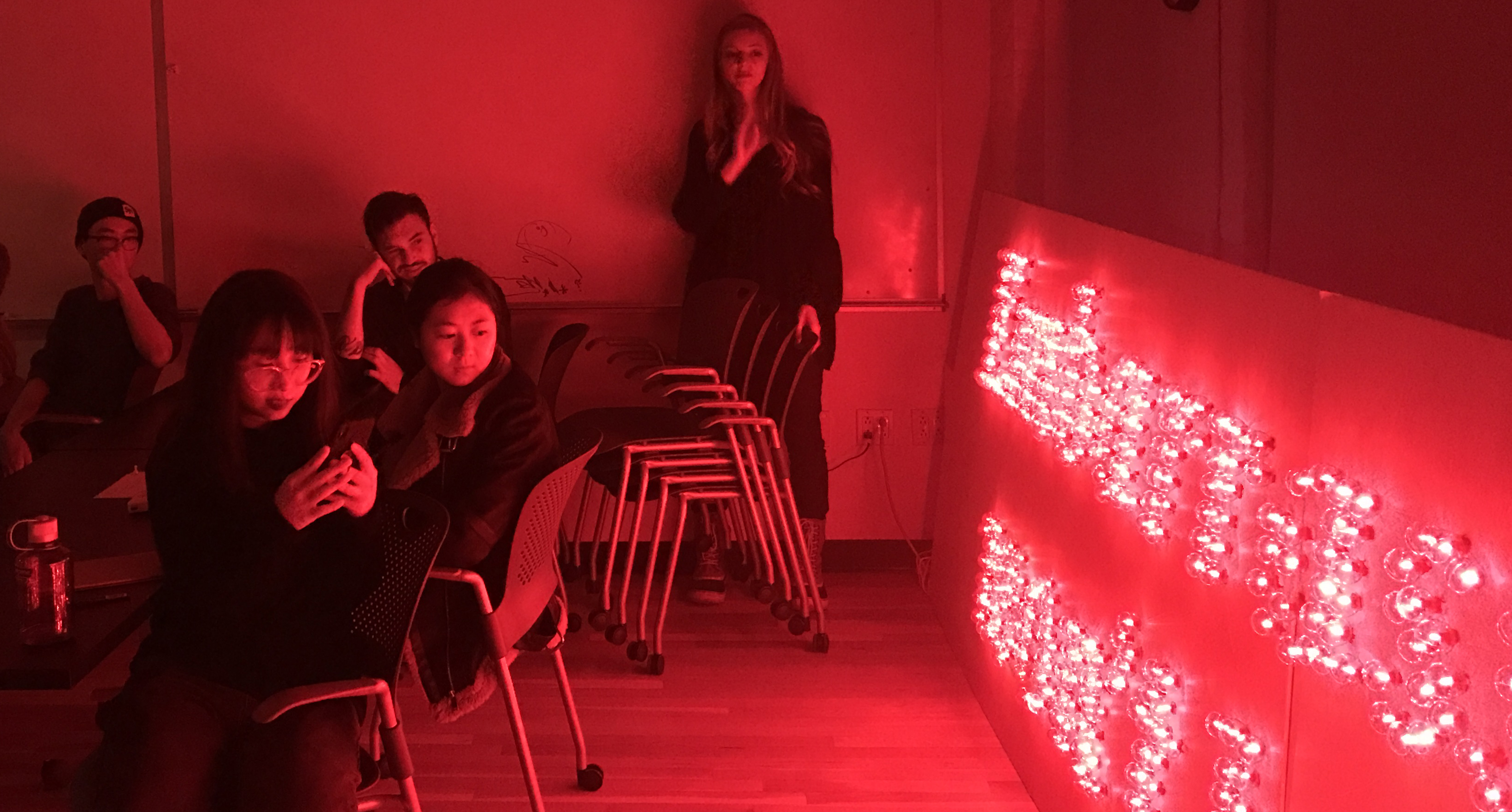 A photo of a group of students looking at a red glowing board that has some text on it made from LEDs.
