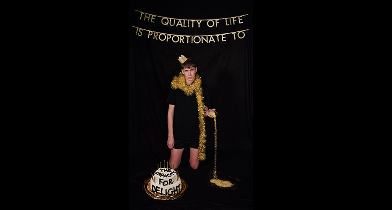 A photo of a person wearing a black dress and a gold tinsel, sitting near a white cake with text on it. On top there is some other text that says: The Quality Of Life is Proportionate To.