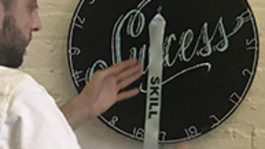 A photo of a person dressed in white and working on some sort of disk object with numbers and the text Success on it. Also on the disk there is a ribbon with the text Skill on it.