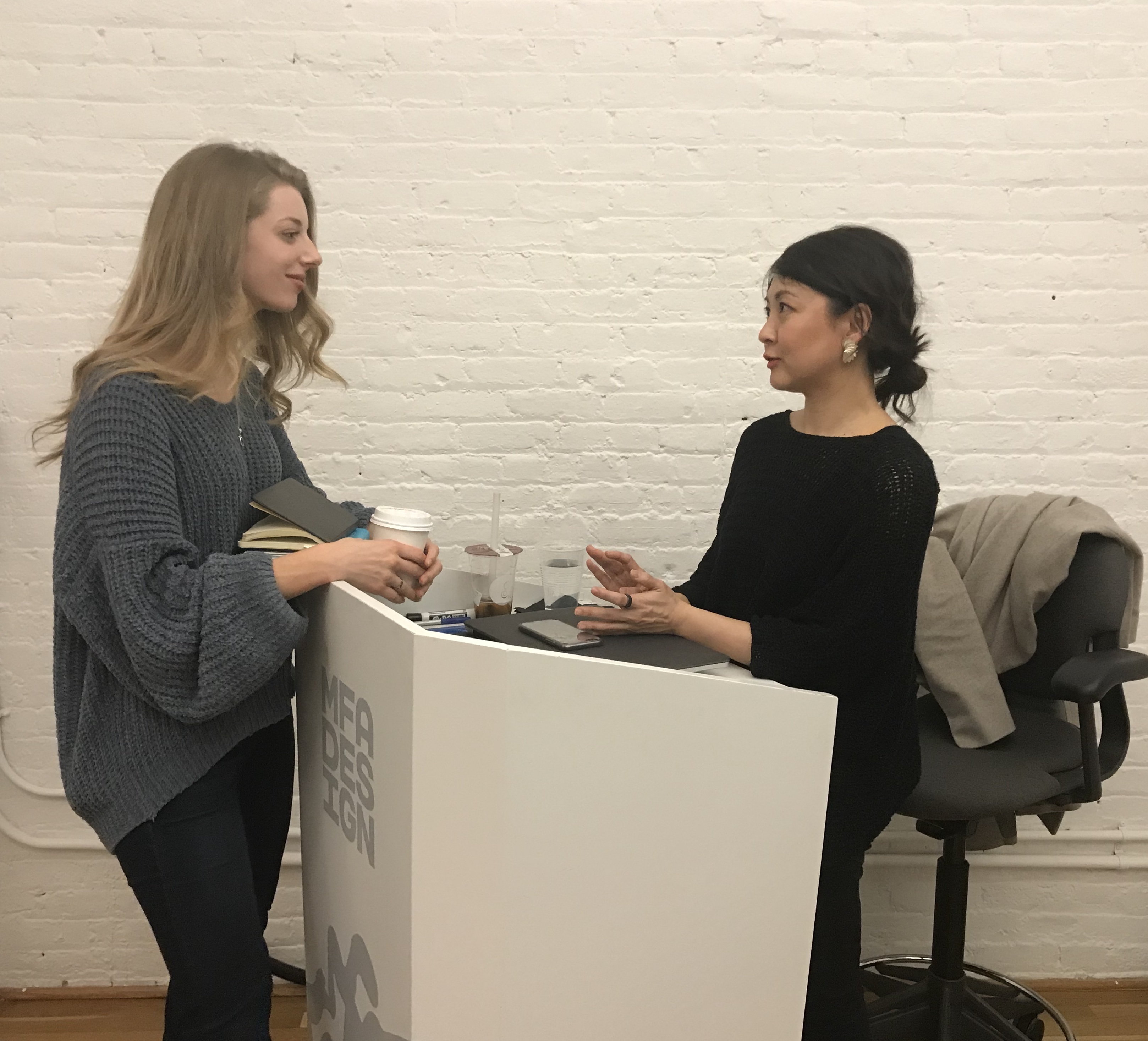 A photo of two women talking. One of them is sitting at a white MFA Design stand.