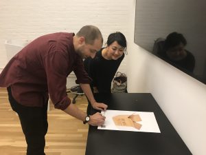 A photo of two people looking at a poster on a table. The poster has an image of a cardboard box on it.