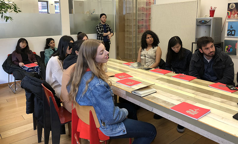 A group of students sitting around a table with some books on it, and listening to someone giving a lecture.
