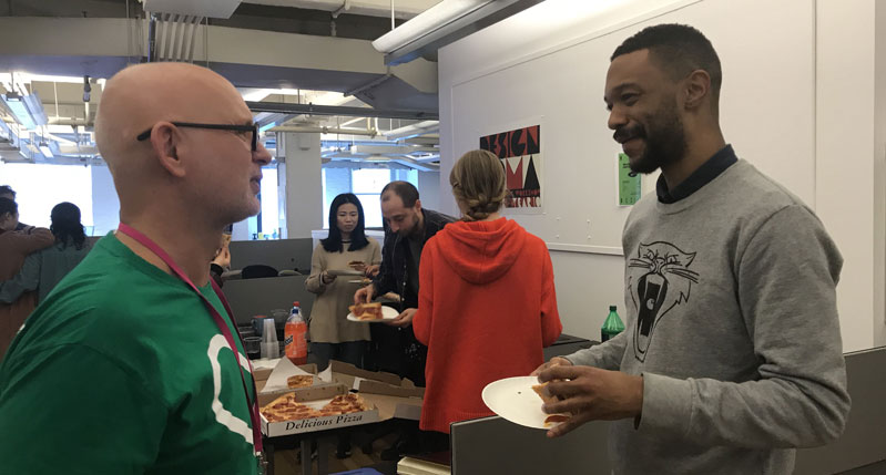 A photo of two people talking while, other people are eating some pizza and drinking juice.