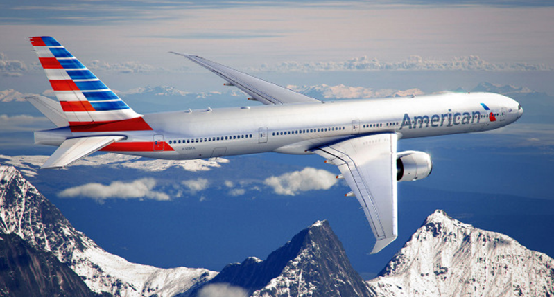 A photo of a silver plane above the mountains and clouds. On its tail and nose there is a US flag and the logo American Airlines.