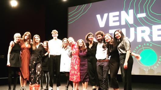 A photo of a group of people sitting on a stage in front of a screen projector that has on it the text: Ven-Ture.