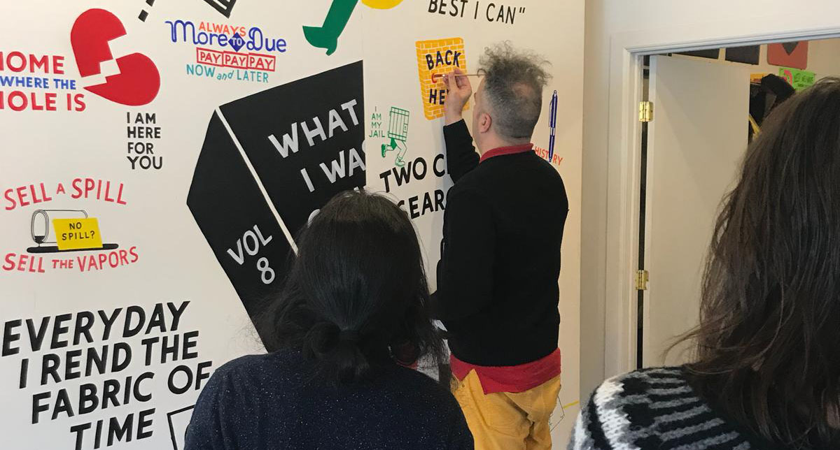 A photo of a man painting different logos on a white wall while other people are watching him.
