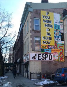 A street view of a building on which there are different artistic banners. On one of them there is the text: If you were here I'd be home now. New York Best ESPO.
