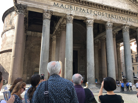 A photo of some people visiting and looking at an old roman building, with some text engraving and pillars at the entrance.