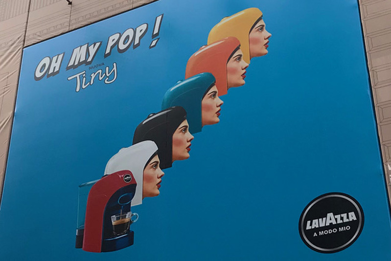 A blue poster showing a 3d computer generated human heads with colorful helmets that look like an espresso machine cover. There is also an espresso machine and the text: Oh my pop! Tiny Lavazza A modo mio..