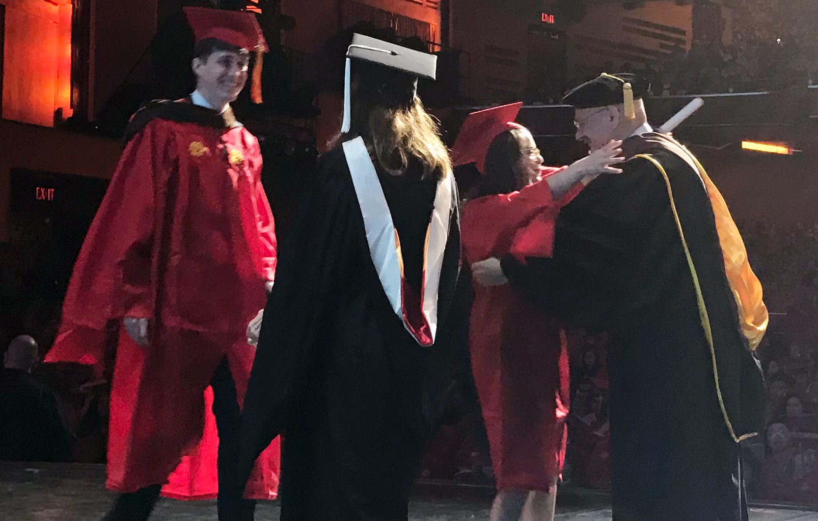 A photo of a group of students and teacher on a stage at the graduation ceremony. They are wearing black or red robes and hats.
