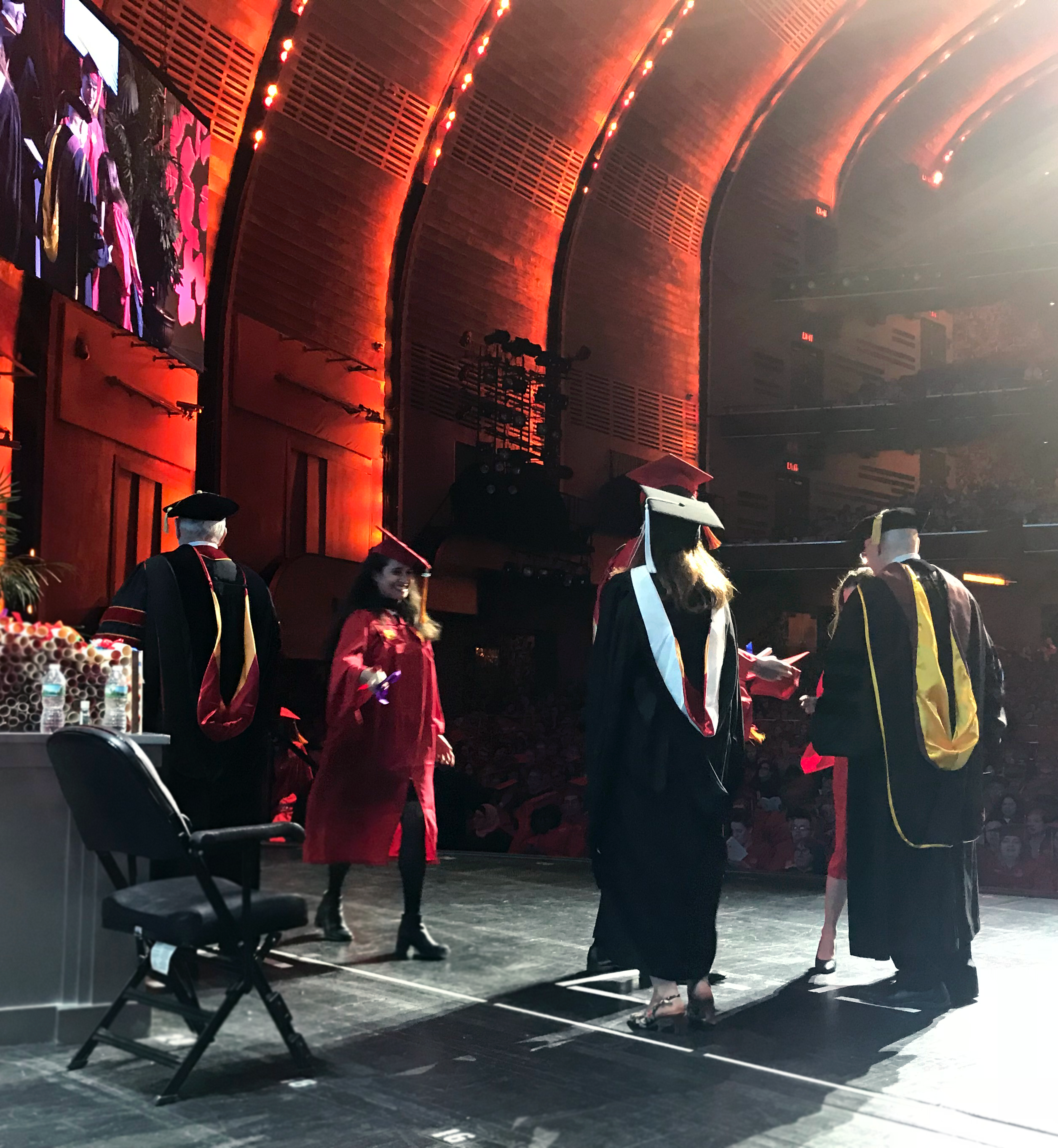 A photo of a group of students and teacher on a stage at the graduation ceremony. They are wearing black or red robes and hats.