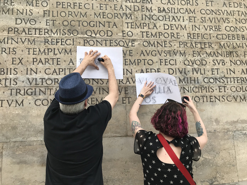 A group of people copying on pieces of paper the letters engraved on a stone wall.