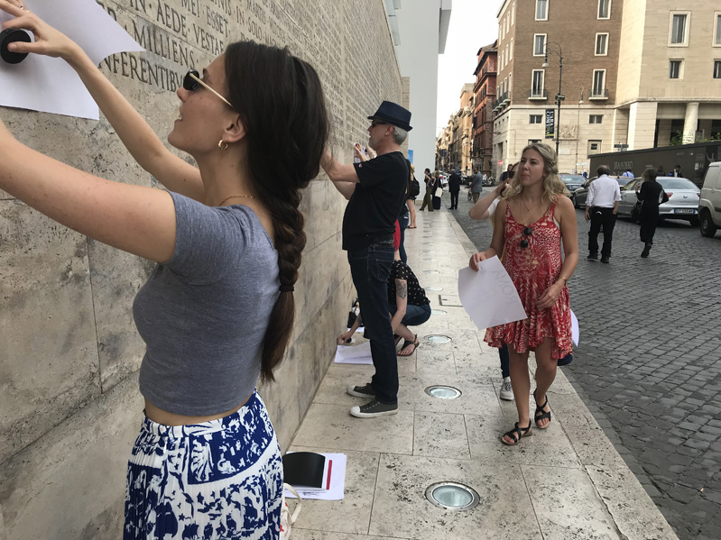 A group of people copying on pieces of paper the letters engraved on a stone wall.