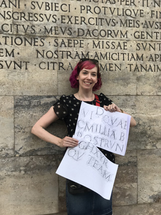 A photo of a girl holding a piece of paper with letters copied from a stone carved wall behind her.