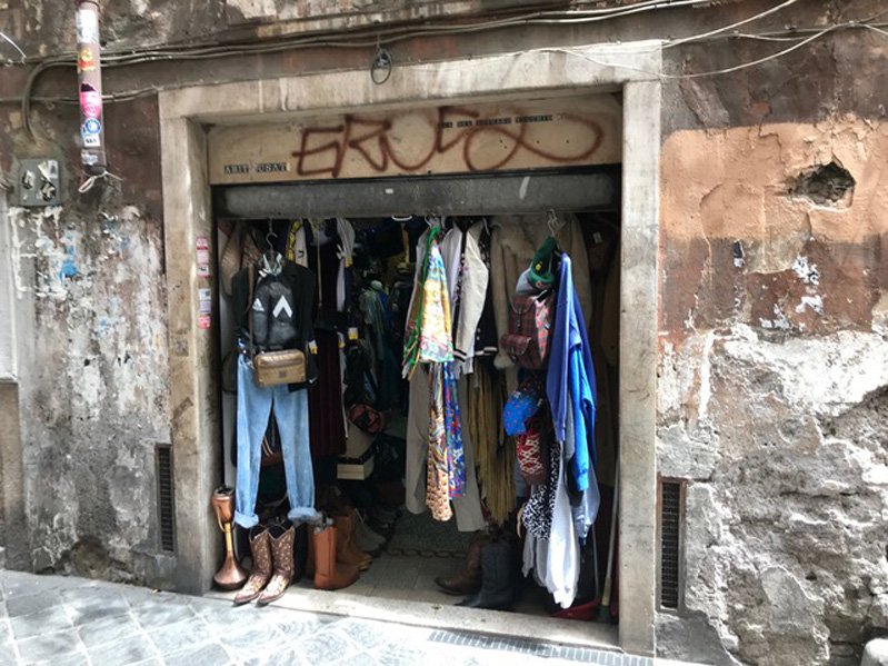 A photo of a store's entrance, made from stone, where there are cloths hanging and some shoes on the sides.