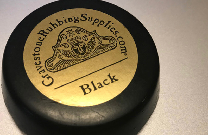 A photo of a black disc with a golden label on which there is a head logo and the text: GravestoneRubbingSuppliers.com. Black.