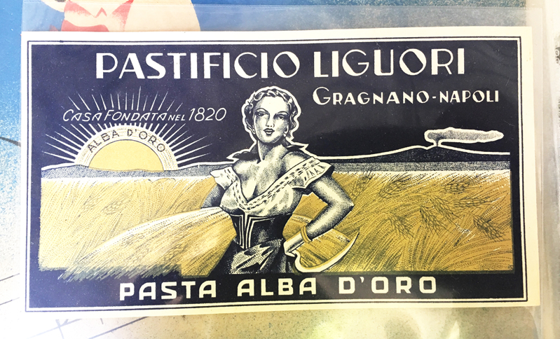 A photo of an old box with a blue and yellow drawing of a lady in a wheat field, while the sun is shining. On it there is the text: Pastificio Liguori Pasta Alba D'Oro.