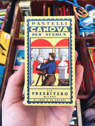 A photo of a hand holding a yellow crayon box with drawings of a man doing a sculpture. On the box there is a text: Pastelli Canova Per Scuola. Presbitero Milano 12 Colori.