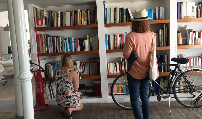 A photo of two girls checking a bookshelf in a room. There is also a bicycle near the shelf.