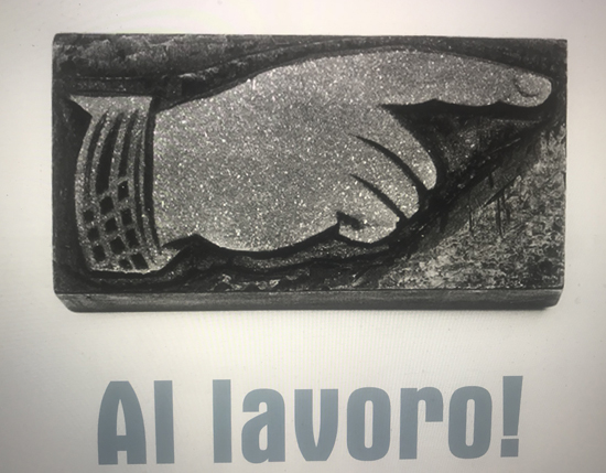 A photo of a 3d stone engraved with a hand that is pointing a finger. There is also the text: Al lavaro!