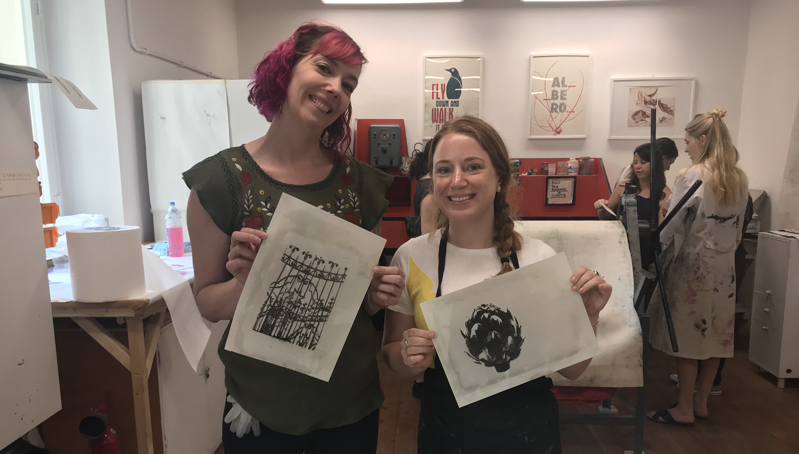 A photo of two women showing their ink printed designs on paper.