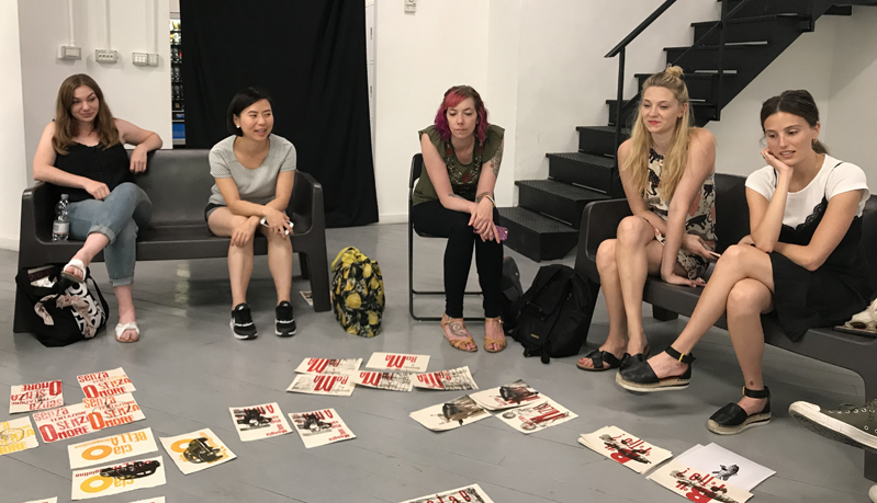 A photo of a group of people sitting on chairs in a circle manner, while looking at some cover book artwork set on the floor in the middle.