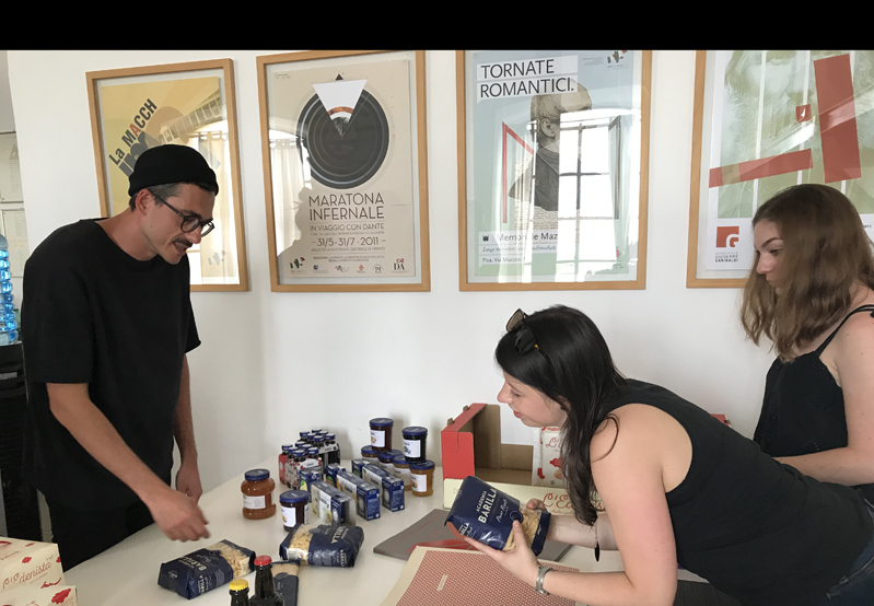 A photo of three people looking at some blue label design on a set of products put on a table. Also near them, hanged on a wall, are a set of posters encased in glass with a wood frame.