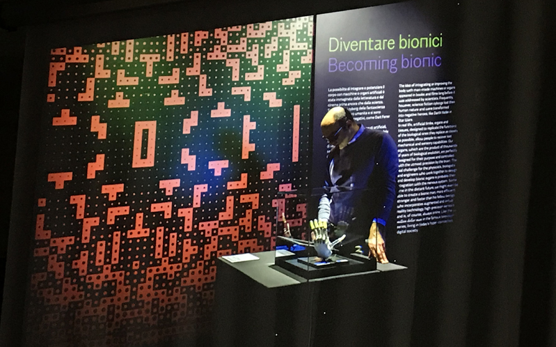 A photo from an art gallery where on the wall is some sort of tetris design and some text with title: Dieventare Bionici Becoming Bionic. Also on a desk is some sort of bionic hand and near it a human figure with a mask.