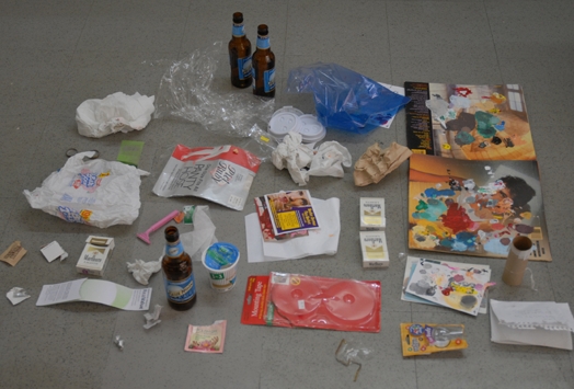 An image depicting cigarette packs, different plastic bottles and bags, razors, papers with paint on them, paper bags, cd shaped empty case and other remains.
