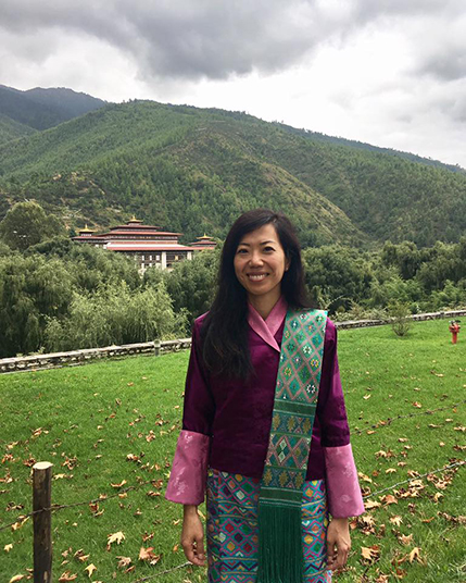 A photo of a woman wearing a pink and green traditional costume while standing in a greenfield and in the background having a mountain with a forest and a temple.