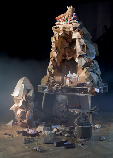 An art setup of what seems like a wooden polygonal shaped cave and near it a wooden polygonal shaped human figure sitting. Around them there are different objects like plastic bottles, a pot with a flower, cables and other garbage looking objects.