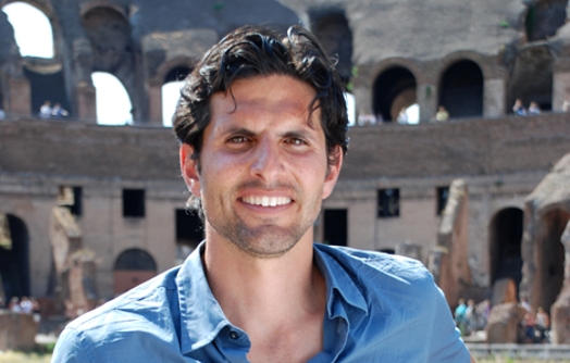 A photo of a man in a blue shirt and in the background the colosseum in Rome.