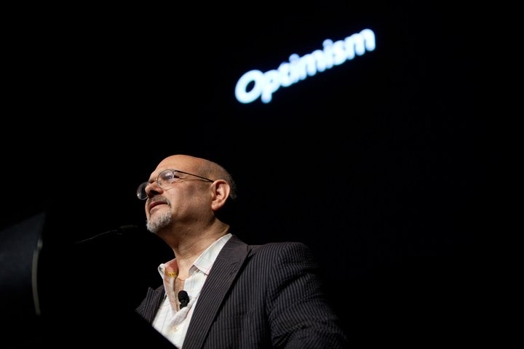 A photo of a man giving a lecture while the text Optimism in blue and white is lit behind him.
